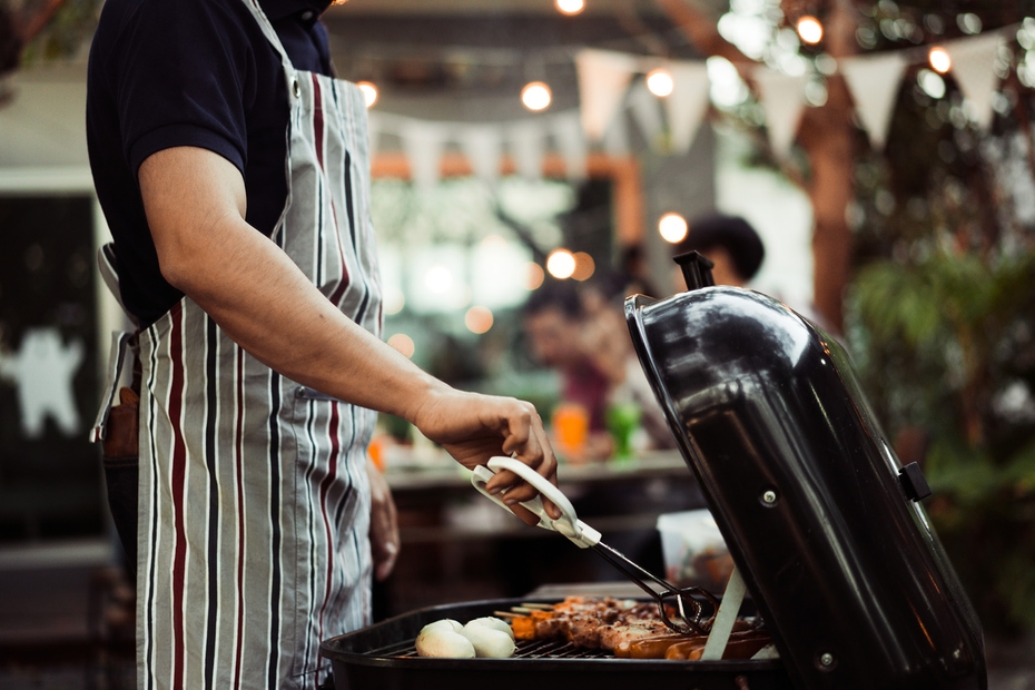 An individual tends to meat on the BBQ. In the background are several students gathered with a family to enjoy dinner outdoors.